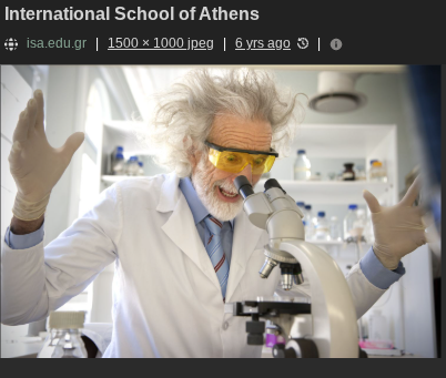 a man with white hair looking into a microscope. he also has a white lab coat and a blue shirt under the lab coat. oh, and a beard. can't forget about the beard. 